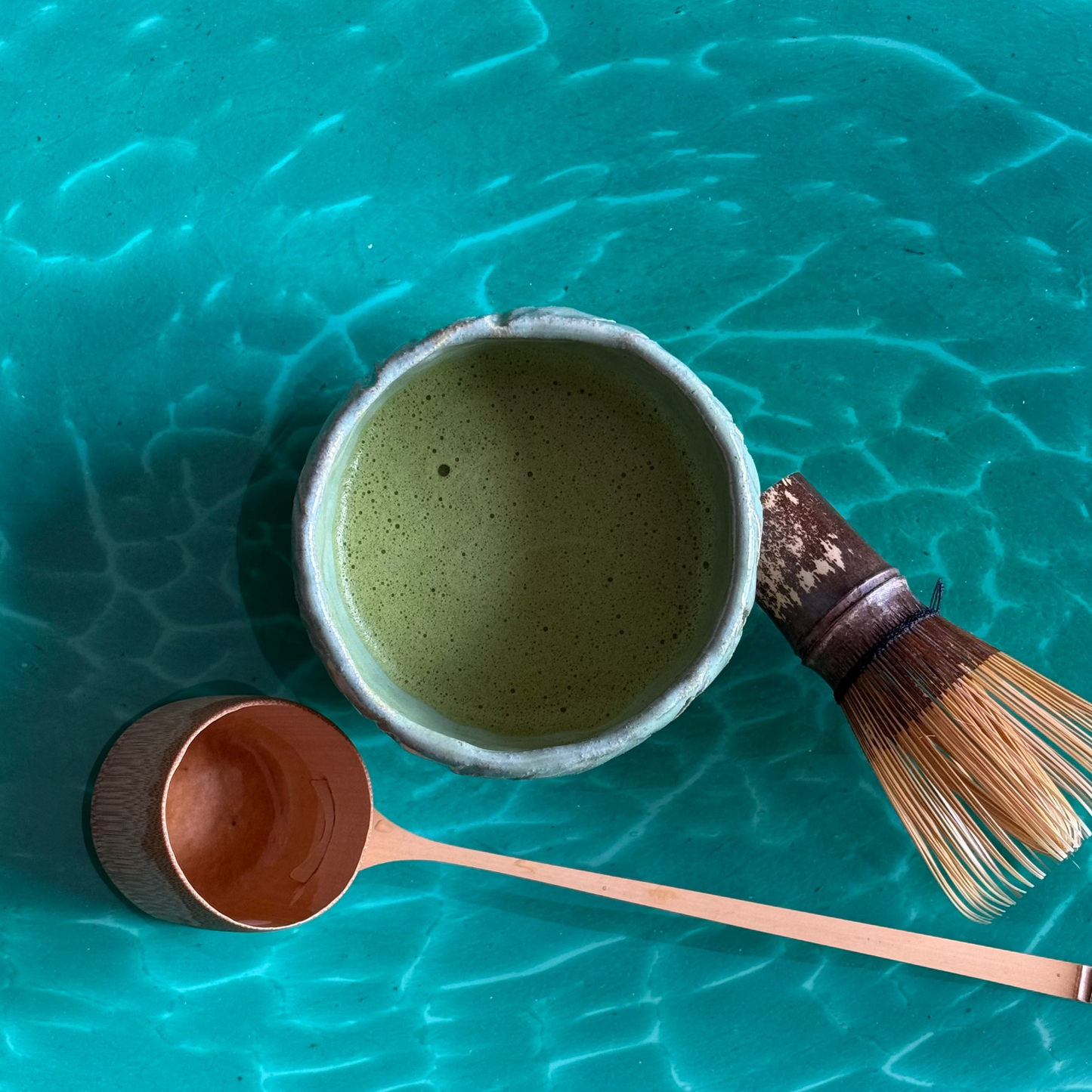 Ceramic Matcha bowl with Matcha contents, Bamboo ladle, and whisk on a teal ocean textured surface