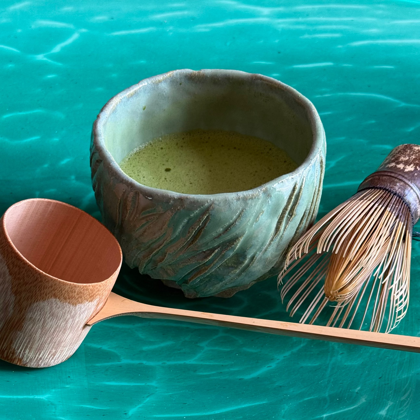 Ceramic bowl with Matcha, Bamboo Ladle, and whisk on a teal ocean background